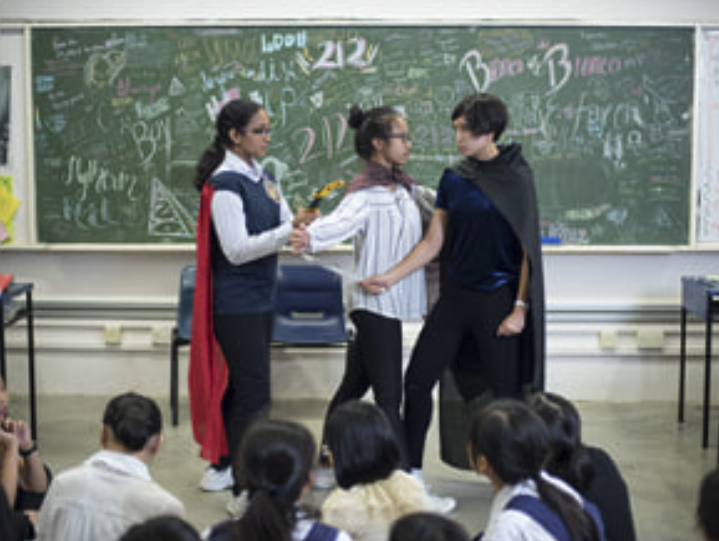 Three students engaged in a theatrical play within a classroom, with other students seated on the floor watching them as audiences.